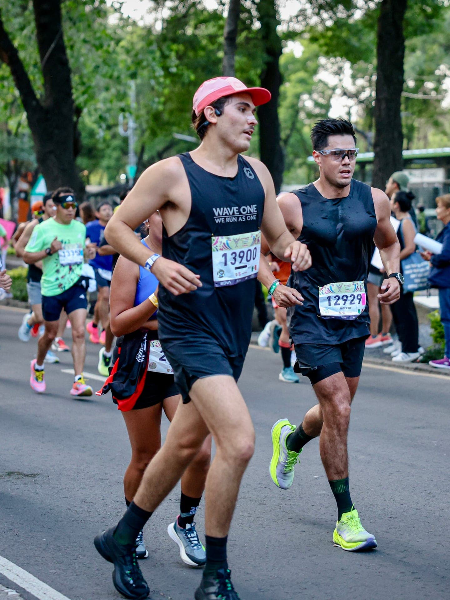 Two Wave One runners in black singlets pacing each other through a tree-lined CDMX race course