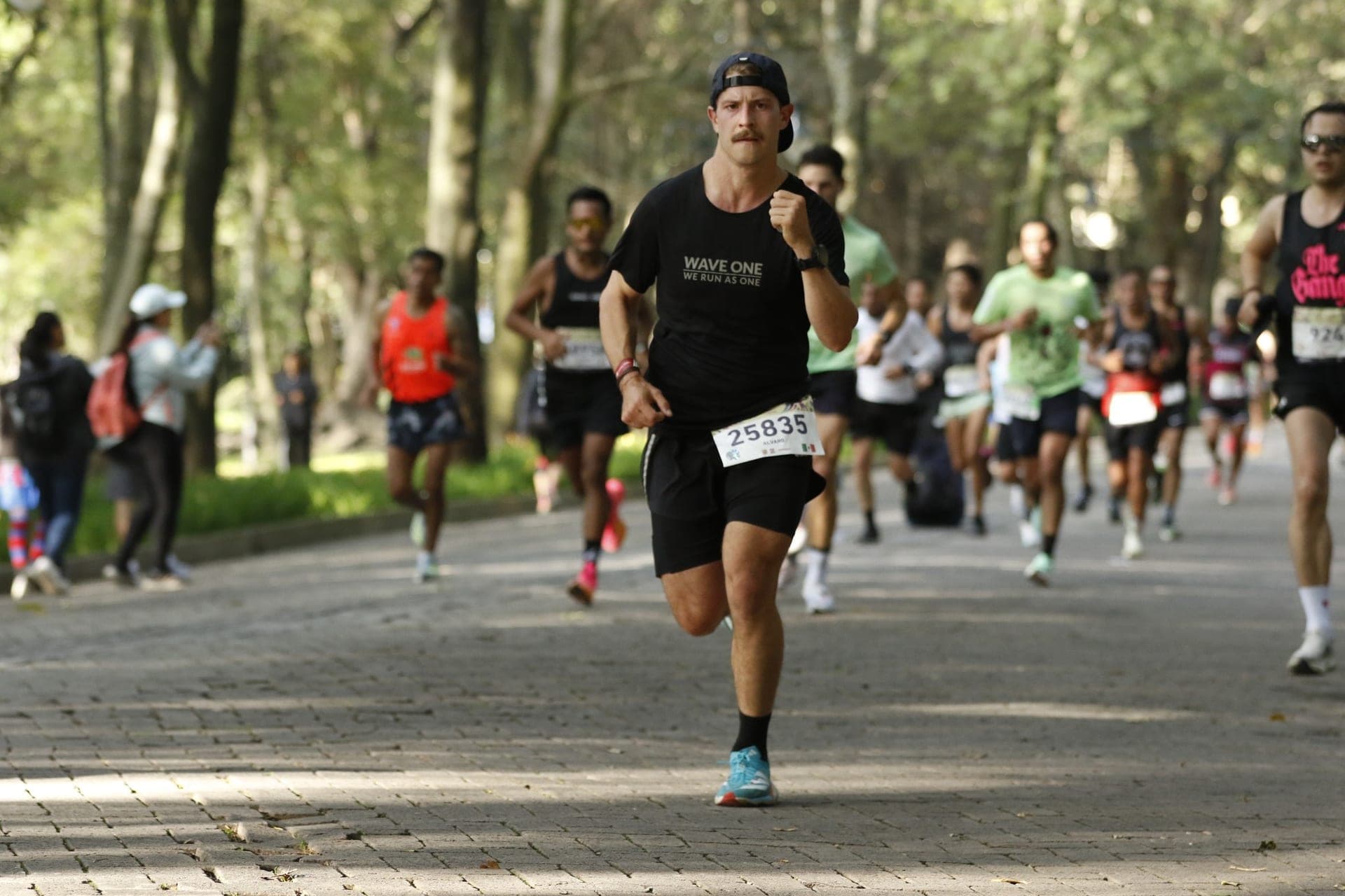 Wave One runner leading a pack along a tree-shaded CDMX park road