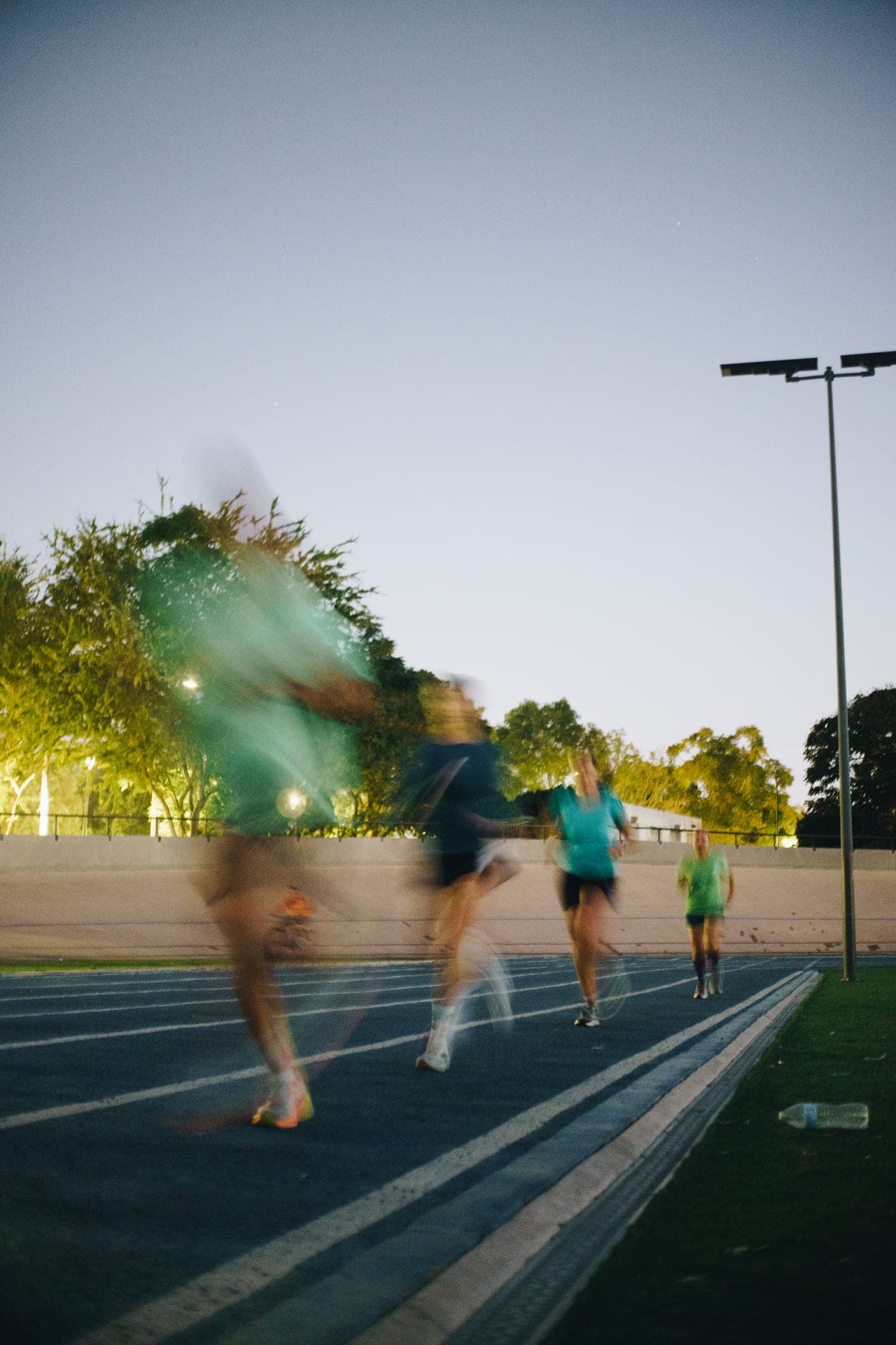 Long-exposure track session at twilight, runners dissolving into colored streaks under a single stadium light