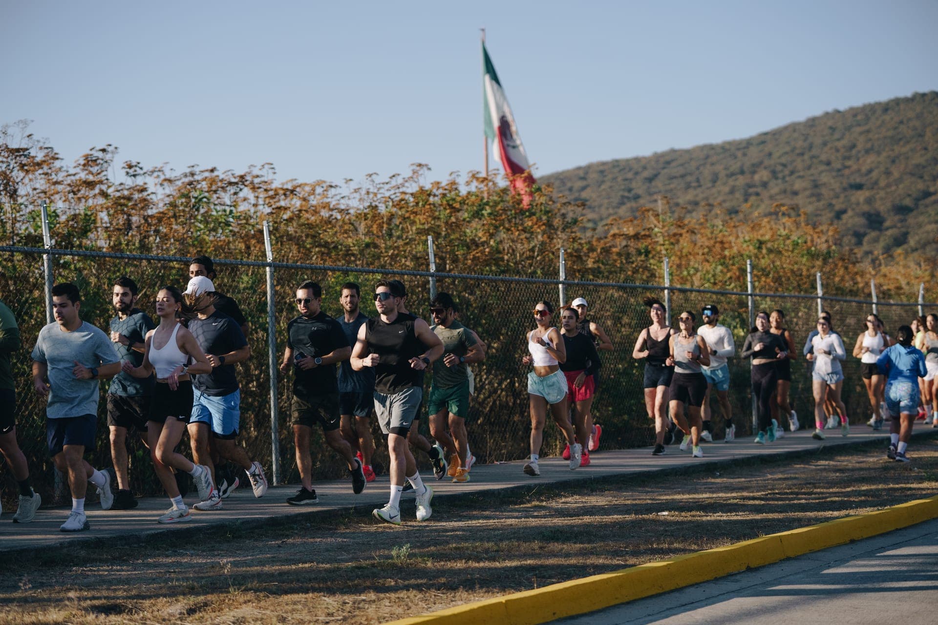 Wave One runners moving along a fenced path with a Mexican flag and mountains behind them