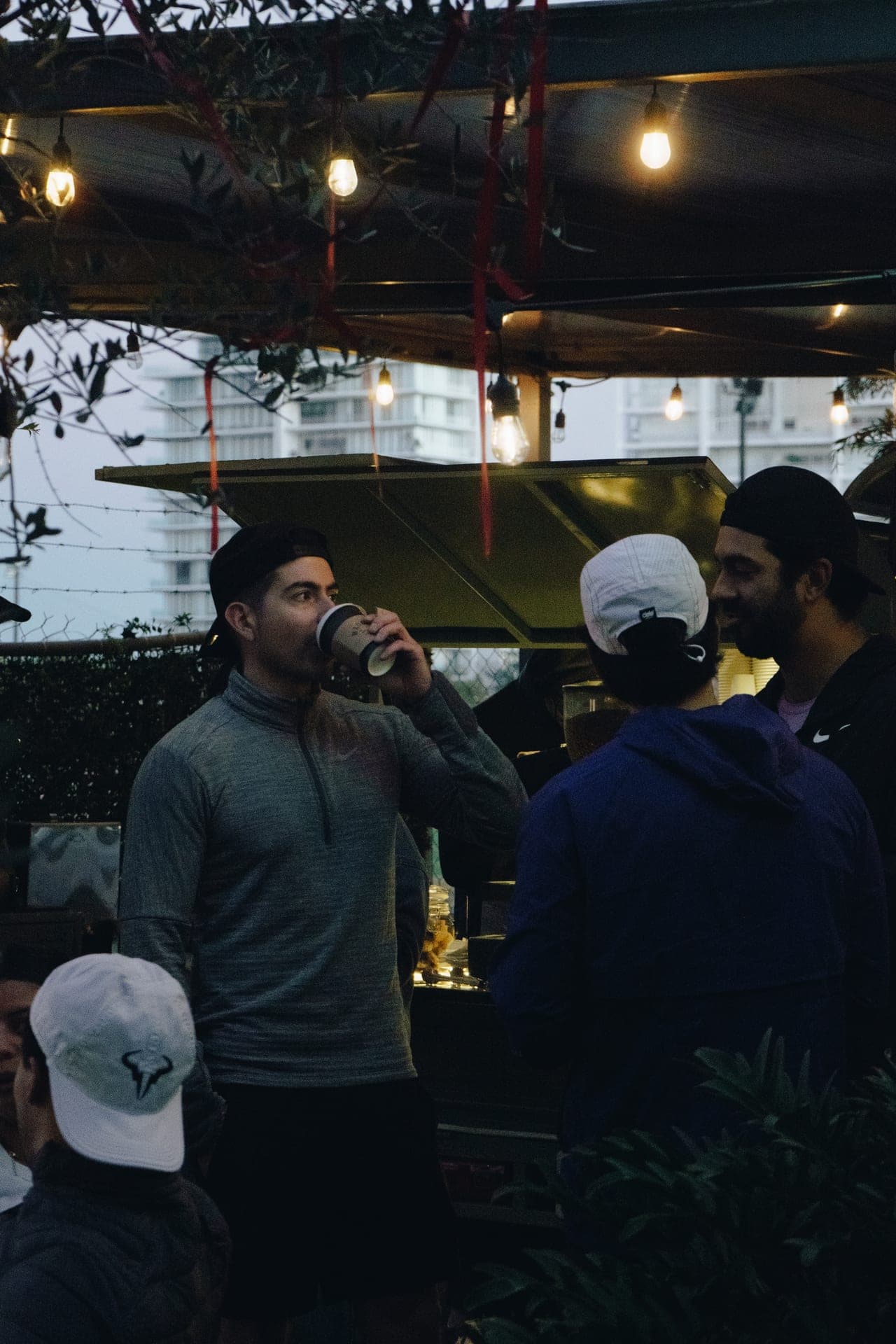 Waver in a grey quarter-zip sipping coffee under string lights at a Friday-morning cafe